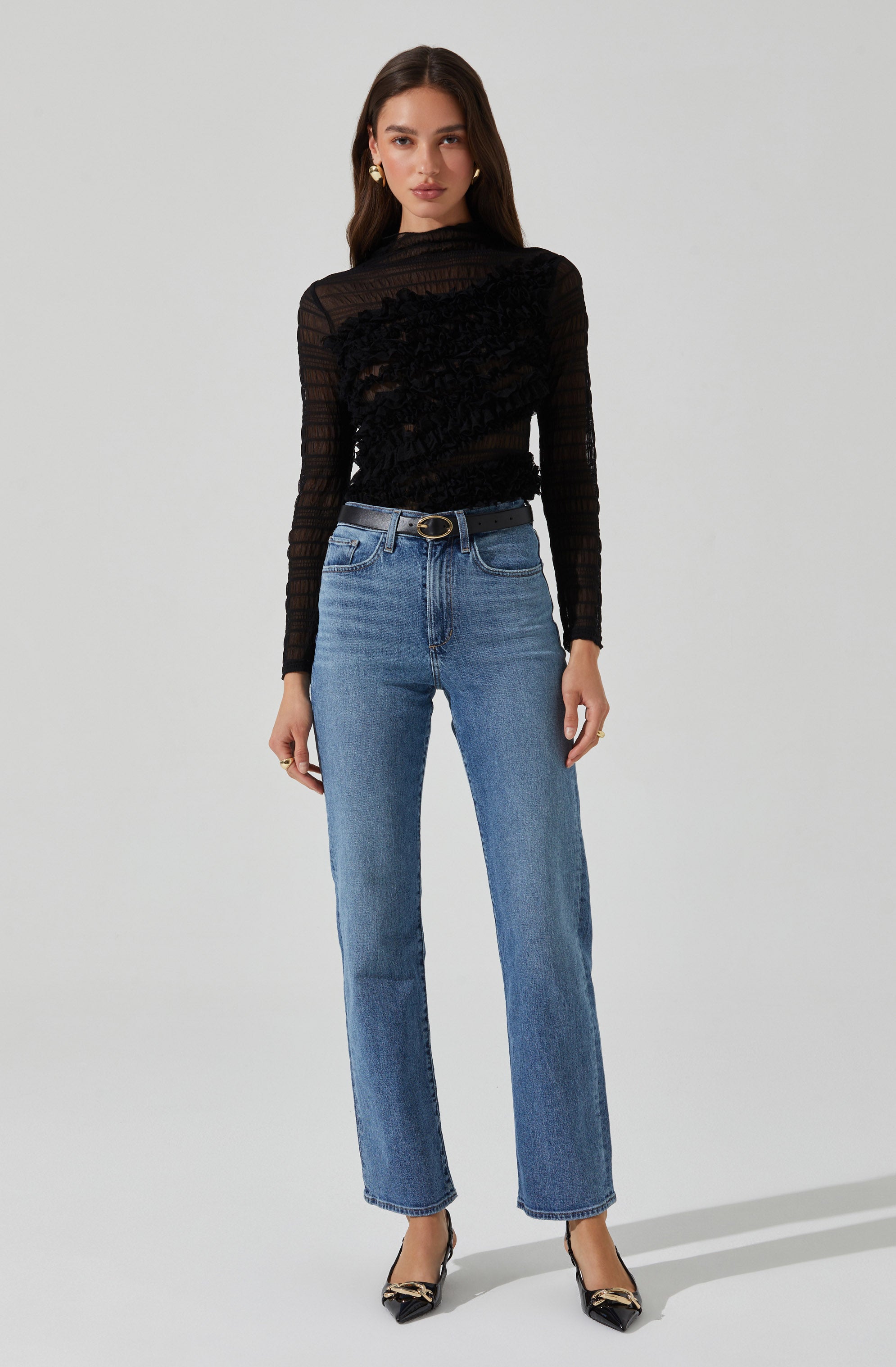 A woman is wearing a sheer black long-sleeve top with horizontal stripe patterns, featuring texture and detailing across the fabric. She pairs it with high-waisted, straight-leg blue jeans that have a classic denim wash. The outfit is accessorized with a black belt with a gold buckle, gold earrings, and pointed black heels with decorative embellishments.