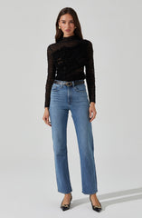 A woman is wearing a sheer black long-sleeve top with horizontal stripe patterns, featuring texture and detailing across the fabric. She pairs it with high-waisted, straight-leg blue jeans that have a classic denim wash. The outfit is accessorized with a black belt with a gold buckle, gold earrings, and pointed black heels with decorative embellishments.