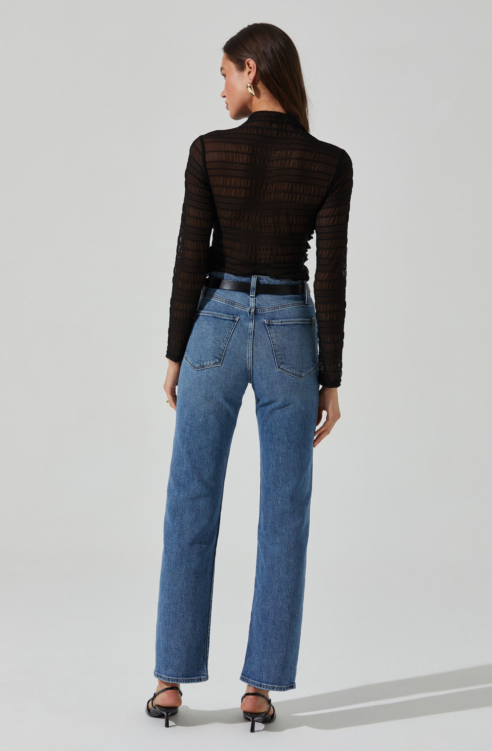 A woman is wearing a black, long-sleeved, sheer top with horizontal striped pattern and a high neckline. She is also wearing high-waisted, loose-fitting blue jeans with back pockets, and black high-heeled sandals.