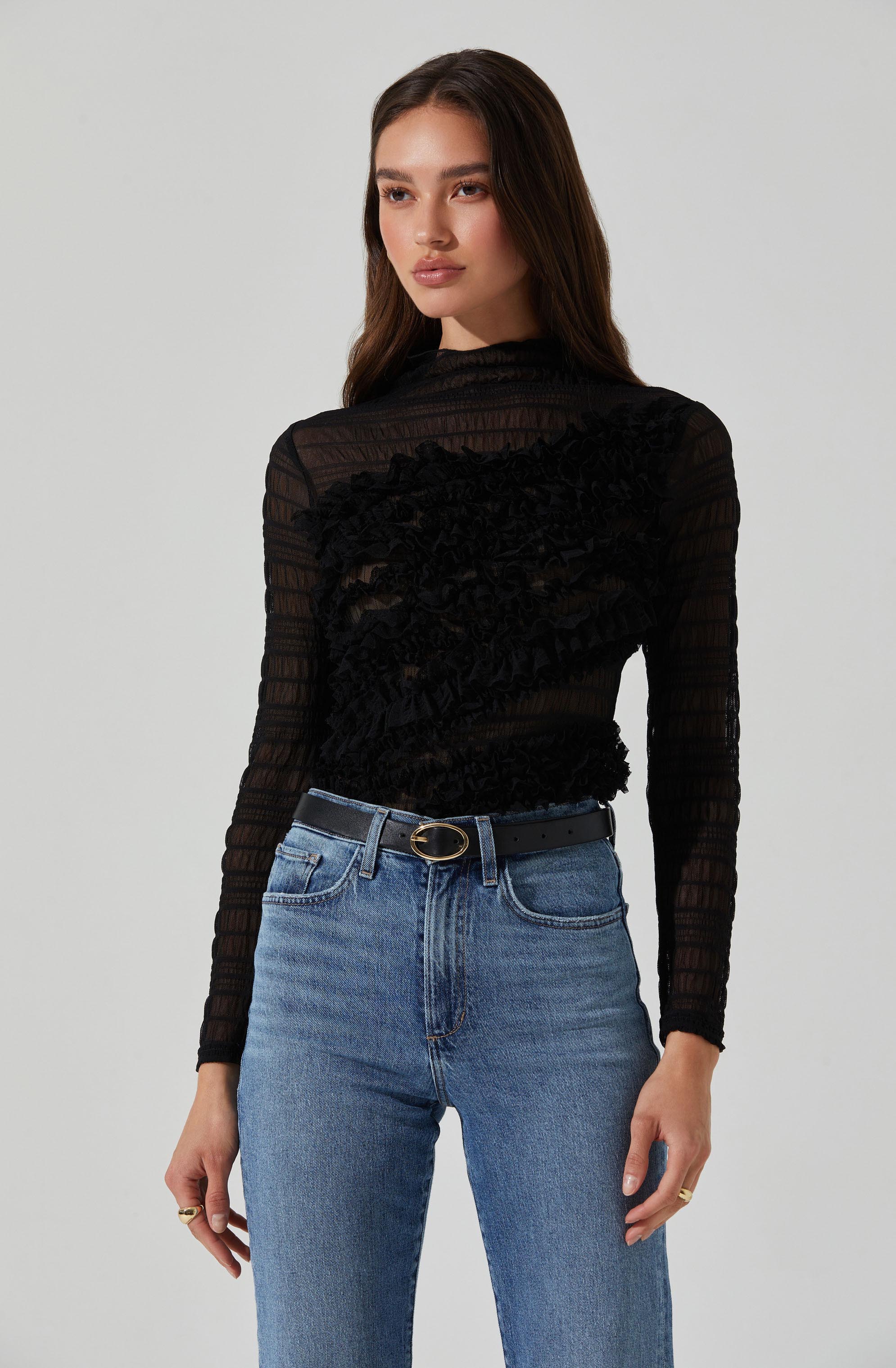 A woman is wearing a black, long-sleeved, semi-sheer top with textured, ruffled detailing across the chest and torso. She pairs the top with high-waisted light blue jeans and a black belt with a gold oval buckle.