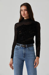 A woman is wearing a black, long-sleeved, semi-sheer top with textured, ruffled detailing across the chest and torso. She pairs the top with high-waisted light blue jeans and a black belt with a gold oval buckle.