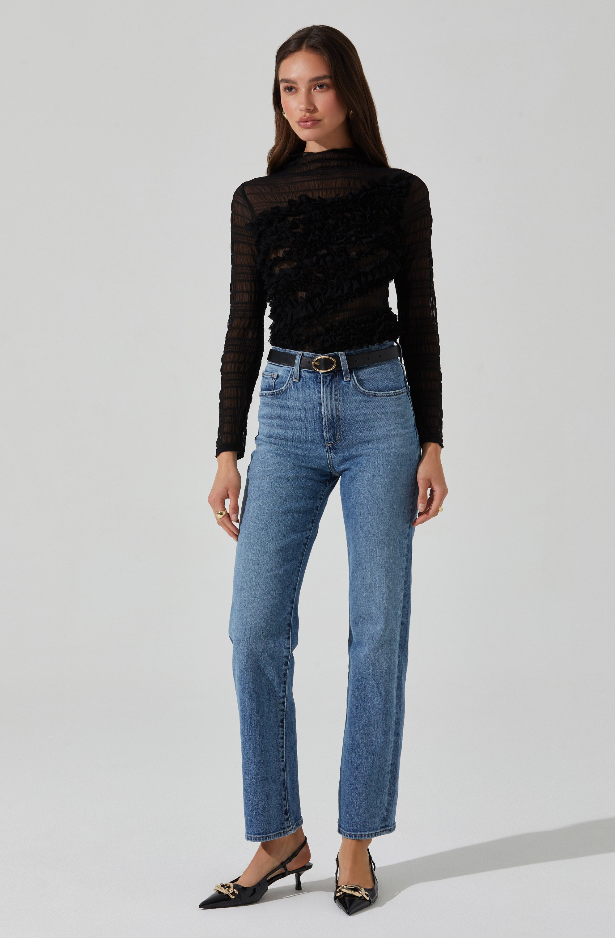 A woman is wearing a black long-sleeve, high-neck, semi-sheer lace top with intricate floral embroidery and textured details. She paired it with high-waisted, straight-leg blue jeans and a black belt with a gold buckle. She is also wearing pointed black high heels with gold decorative accents.