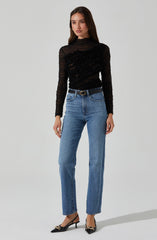 A woman is wearing a black long-sleeve, high-neck, semi-sheer lace top with intricate floral embroidery and textured details. She paired it with high-waisted, straight-leg blue jeans and a black belt with a gold buckle. She is also wearing pointed black high heels with gold decorative accents.