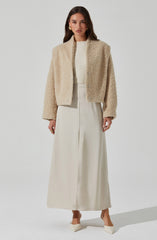A woman is wearing a cream-colored, plush, teddy bear coat with a high collar and buttons down the front. Underneath, she has on a ribbed, cream-colored top. She pairs these with a long, flowing, cream-colored skirt and pointed-toe heels that match her outfit. She also has gold earrings and rings as accessories.