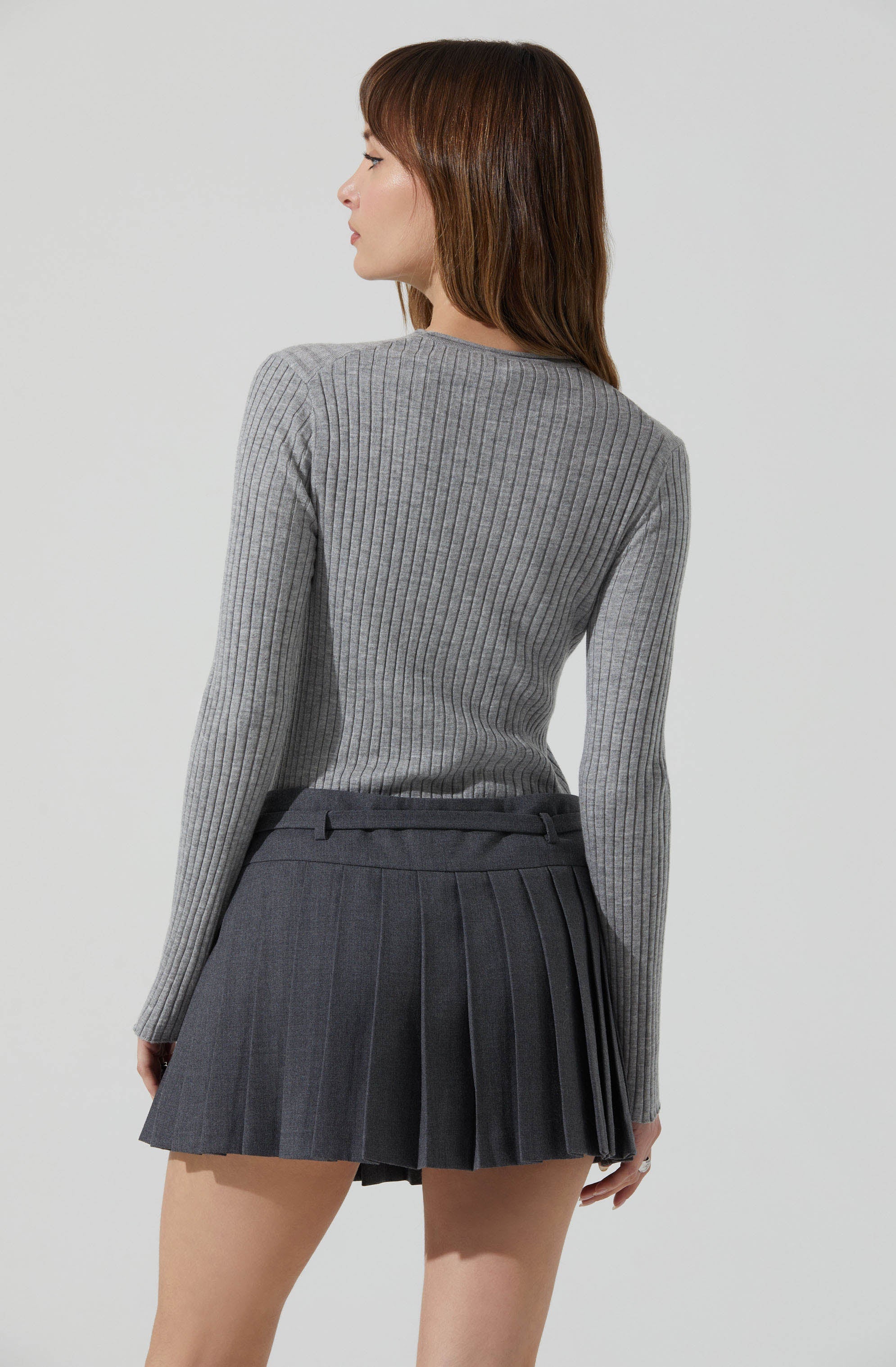 A woman is wearing a fitted, long-sleeve, ribbed, gray sweater with a round neckline, paired with a high-waisted, dark gray pleated mini skirt.