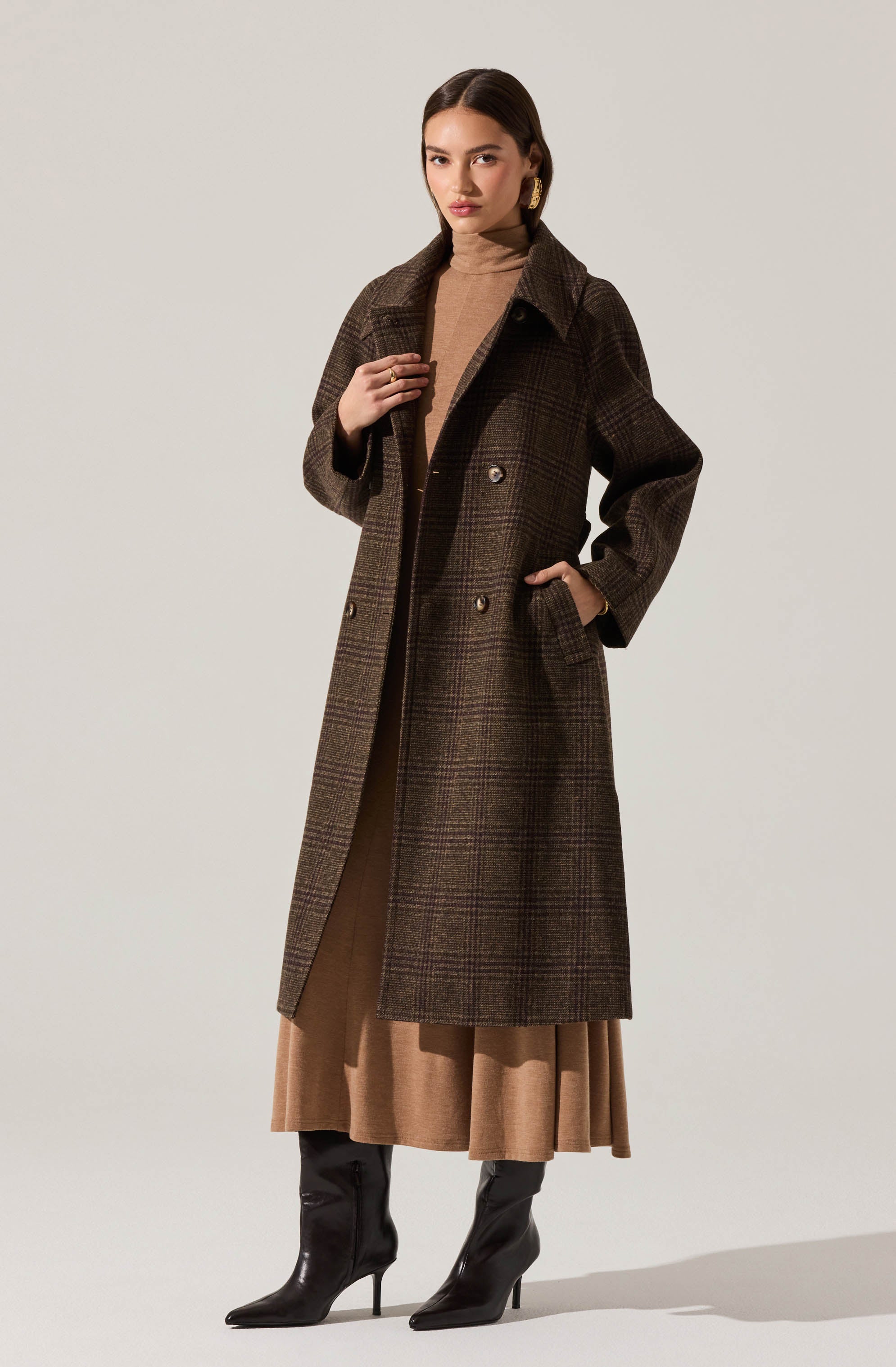 A woman is wearing a long, dark brown, plaid-patterned coat with double-breasted buttons and a wide collar. Underneath, she has on a light brown turtleneck sweater and a matching brown skirt that extends below the coat. She is also wearing black high-heeled boots.