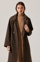 A woman is wearing a long, double-breasted coat with a dark brown and green plaid pattern. Underneath, she has on a high-neck, camel-colored dress or top. She is accessorized with gold hoop earrings and a ring on her left hand.