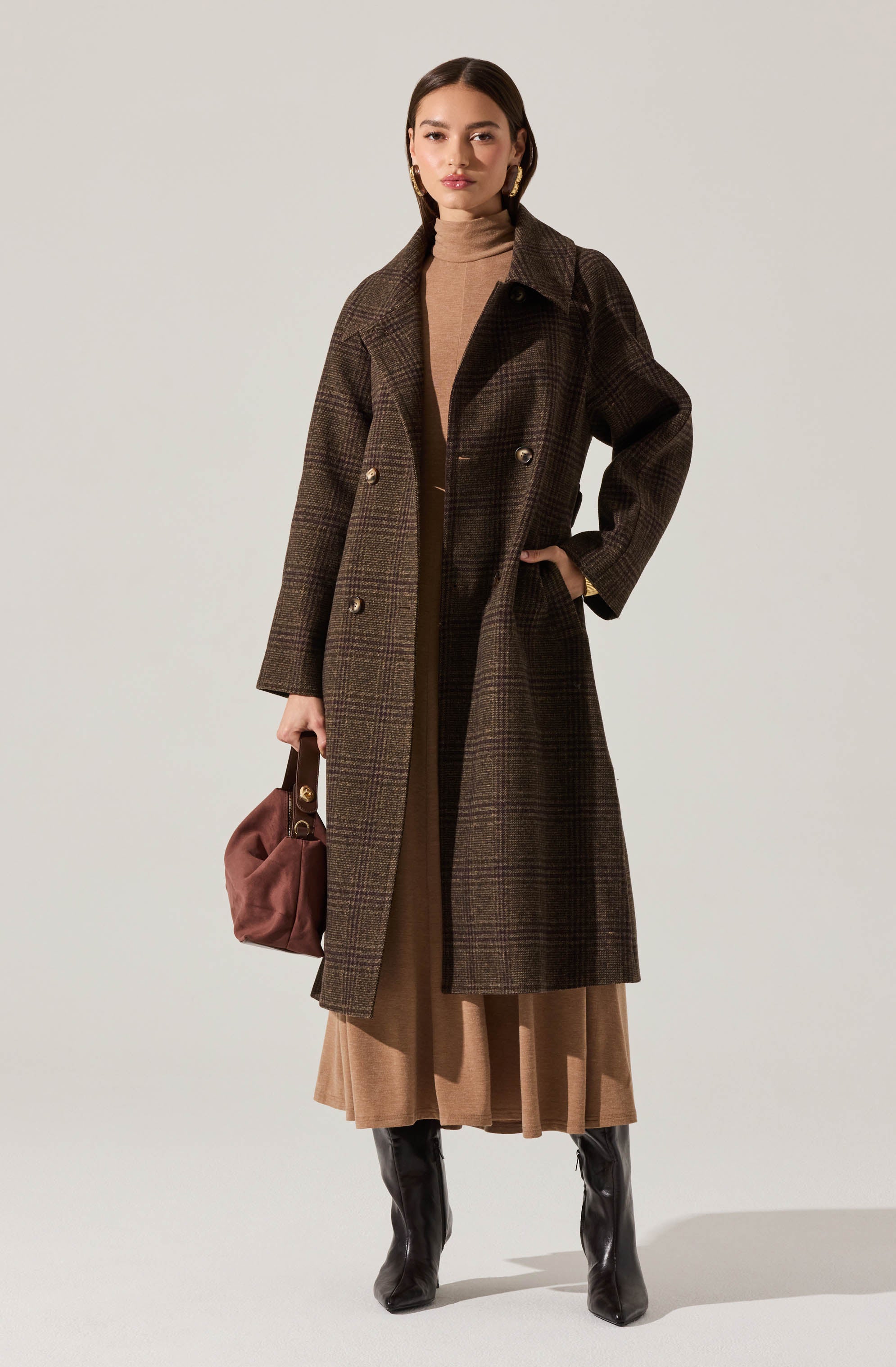 A woman is wearing a long, dark brown plaid coat with a double-breasted design and wide lapels. Underneath, she has on a beige turtleneck sweater. She is also wearing a beige pleated skirt that reaches mid-calf and pairs with black knee-high boots. She holds a small brown handbag with a gold clasp and hardware, and her accessories include large, gold hoop earrings.