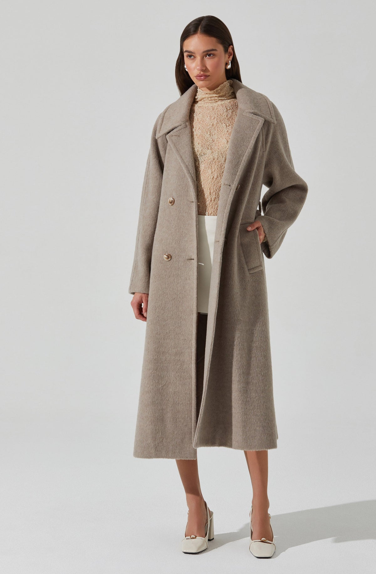 The woman is wearing a long, light beige or taupe-colored wool coat with a wide collar and two buttons, styled open. Underneath, she has on a cream-colored, high-neck, textured sweater. She is also wearing white or cream-colored wide-leg pants and white high-heeled shoes.