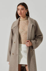 A woman is wearing a long, beige wool coat with large lapels and double-breasted buttons. Underneath, she has on a light, textured high-neck top in a neutral tone, and a white, high-waisted skirt that is above knee length. She has dark, straight hair and is accessorizing with earrings and a ring.