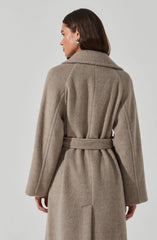 A woman is wearing a long, beige, wool coat with a belted waist. The coat has a wide collar, full-length sleeves, and a slit at the back.