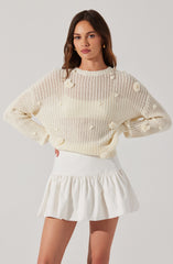 A woman is wearing a cream-colored, chunky knit sweater with a loose fit. The sweater features a distressed design with intentional rips and holes throughout. She pairs it with a white, high-waisted mini skirt that has a gathered, ruffled hem.