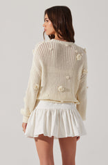 A woman is wearing a cream-colored, oversized knit sweater with textured pom-pom details scattered throughout. She pairs it with a high-waisted, white, ruffled mini skirt.