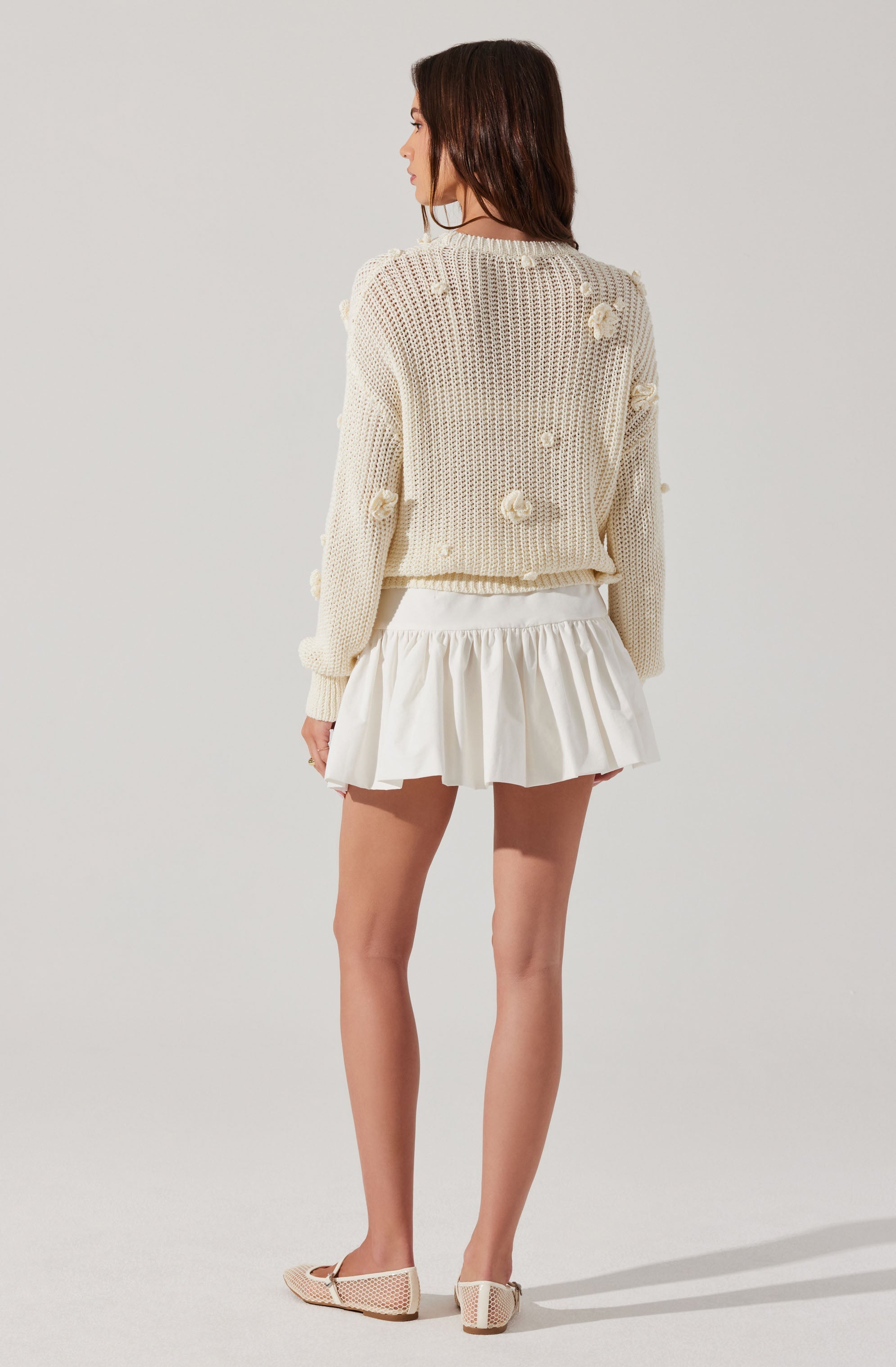 A woman is wearing a cream-colored, chunky knit sweater with textured pom-pom details scattered across the fabric. She pairs it with a white, ruffled short skirt and beige, open-toed shoes with a woven pattern.
