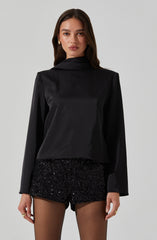 A woman is wearing a long-sleeved black satin blouse with a wide collar, paired with high-waisted black sequin shorts that are fitted and shiny. She also has sheer black tights and gold earrings, and is accessorizing with rings.