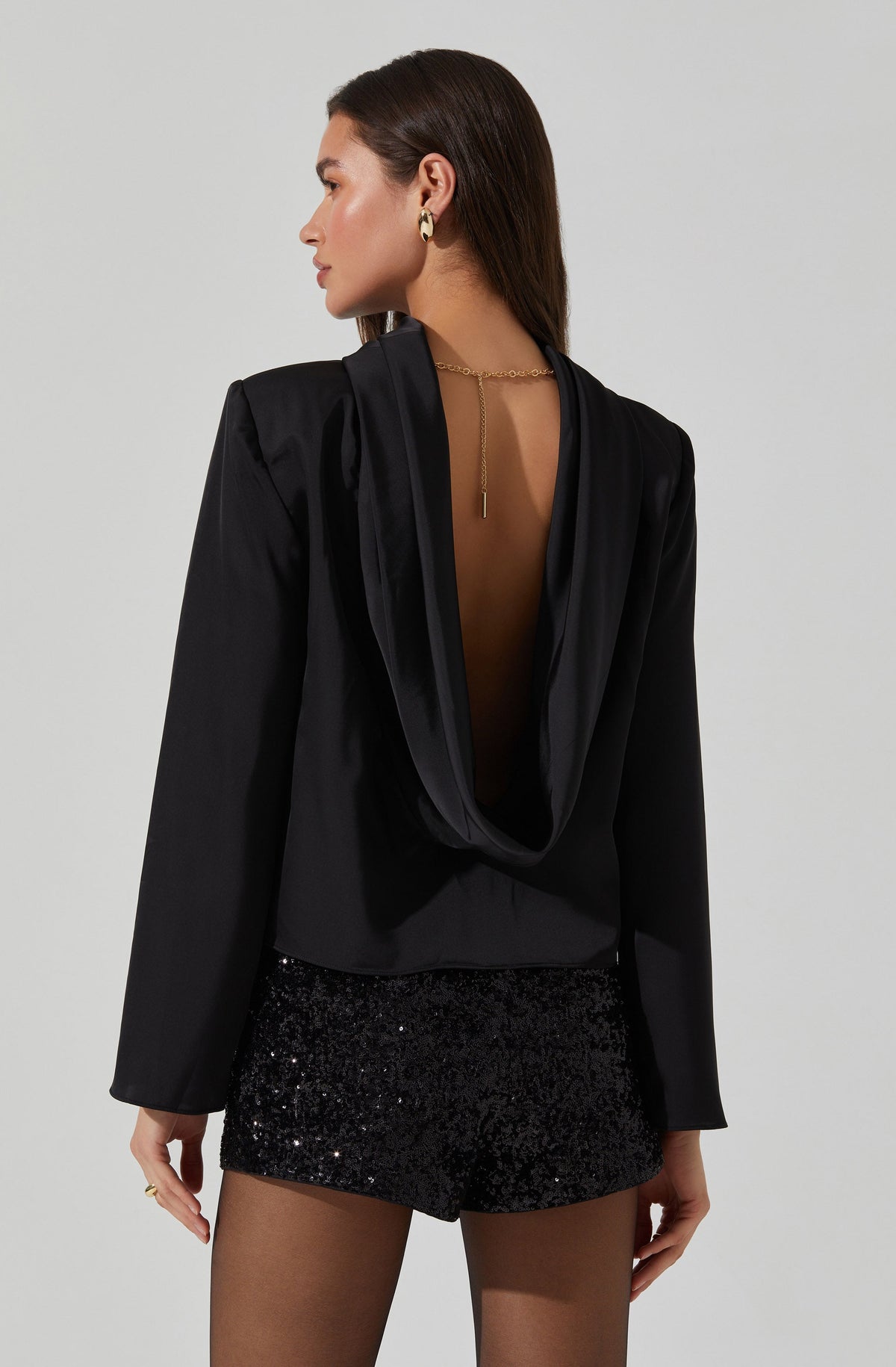 A woman is wearing a black satin blazer with a draped, open-back design. She has paired it with black sequin shorts and is accessorized with gold hoop earrings and a gold chain necklace with a dangling pendant.