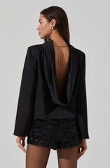 A woman is wearing a black satin blazer with a draped, open-back design. She has paired it with black sequin shorts and is accessorized with gold hoop earrings and a gold chain necklace with a dangling pendant.
