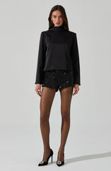A woman is wearing a black, satin long-sleeve top with a high neckline, paired with black, sequined shorts that are short in length. She is also wearing black high-heeled shoes with a decorative embellishment on the toes.