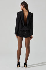 A woman is wearing a black satin blazer with a deep, open back that drapes elegantly, paired with black sequined shorts. She completes the outfit with black high heels and a delicate gold necklace.