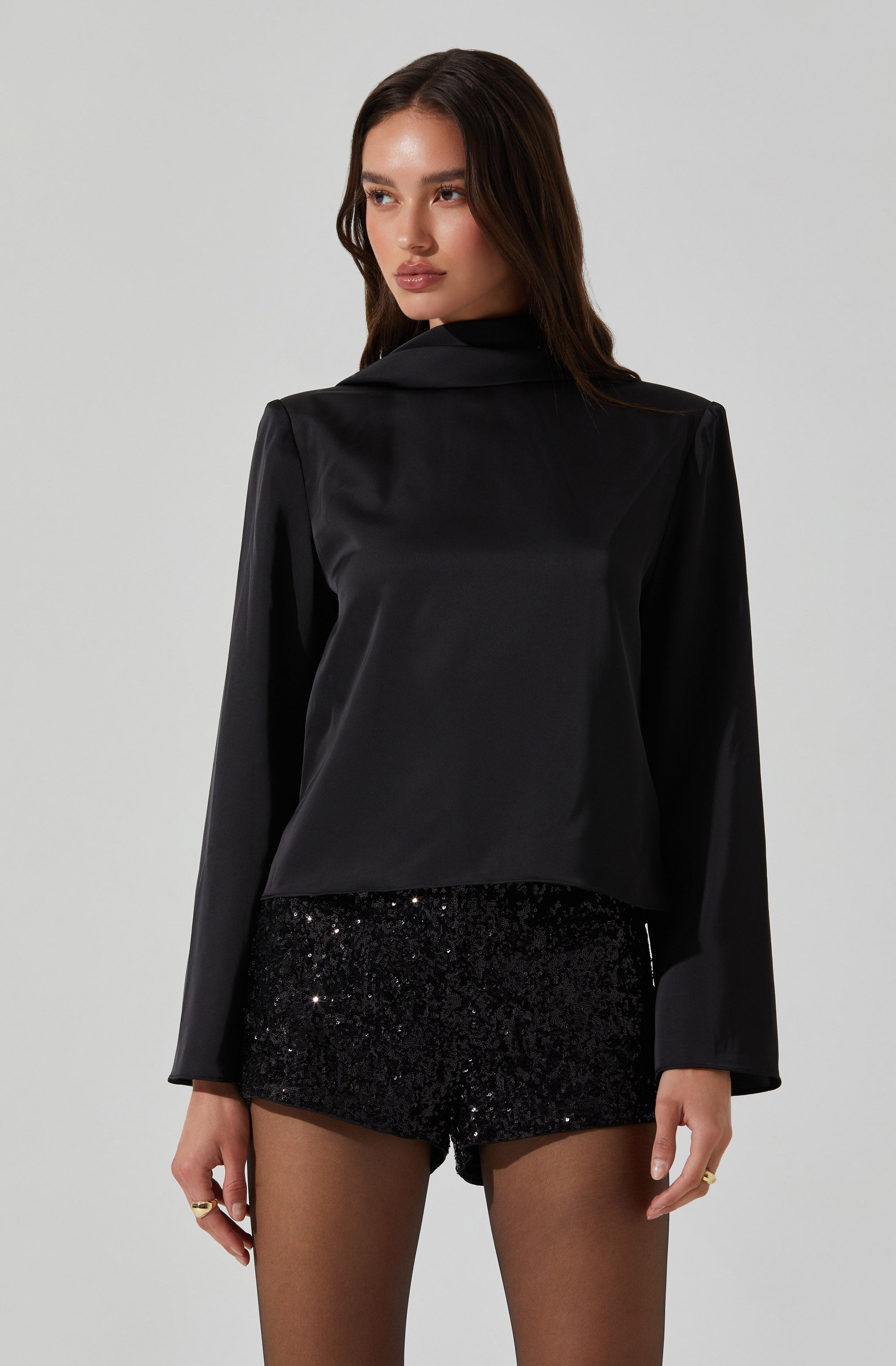A woman wearing a black satin blazer with long sleeves and a notched lapel collar, paired with black sequin shorts, and sheer black tights. She has long, dark hair and is standing against a plain, light-colored background.