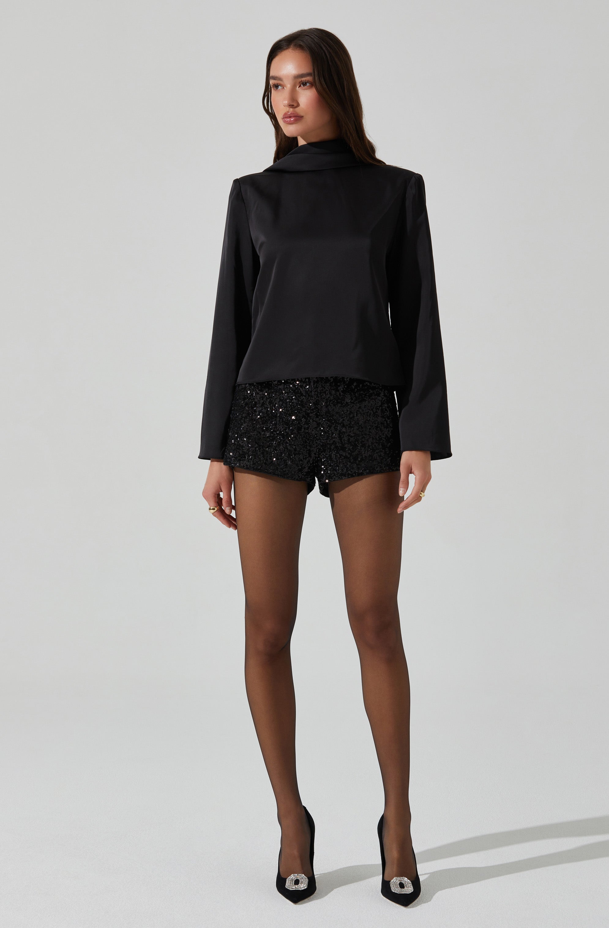 The woman is wearing a long-sleeved, black satin blazer with a notched collar. She has paired it with high-waisted black shorts that are covered in black sequins, creating a sparkling effect. She is also wearing black sheer tights and black high-heeled shoes with a decorative embellishment on the toe.