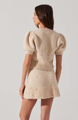 A woman is wearing a beige knit sweater with short puffed sleeves and a crew neckline. The sweater is decorated with small, textured embellishments around the hem and back. She is also wearing a matching beige knit skirt that is decorated with similar textured embellishments.