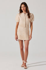 A young woman is wearing a matching cream-colored knit set consisting of a short-sleeved, button-up cardigan with puffed sleeves and a high neckline, paired with a high-waisted, mini skirt featuring a textured pattern with small, decorative embellishments. She completes her outfit with beige ankle-strap shoes.