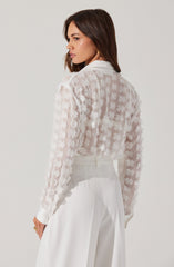 A woman is wearing a sheer, white, long-sleeved blouse with a floral pattern consisting of small, textured flowers. She pairs the blouse with high-waisted, white wide-leg pants. The overall look is elegant and monochromatic, with the blouse featuring intricate detailing of flower motifs.