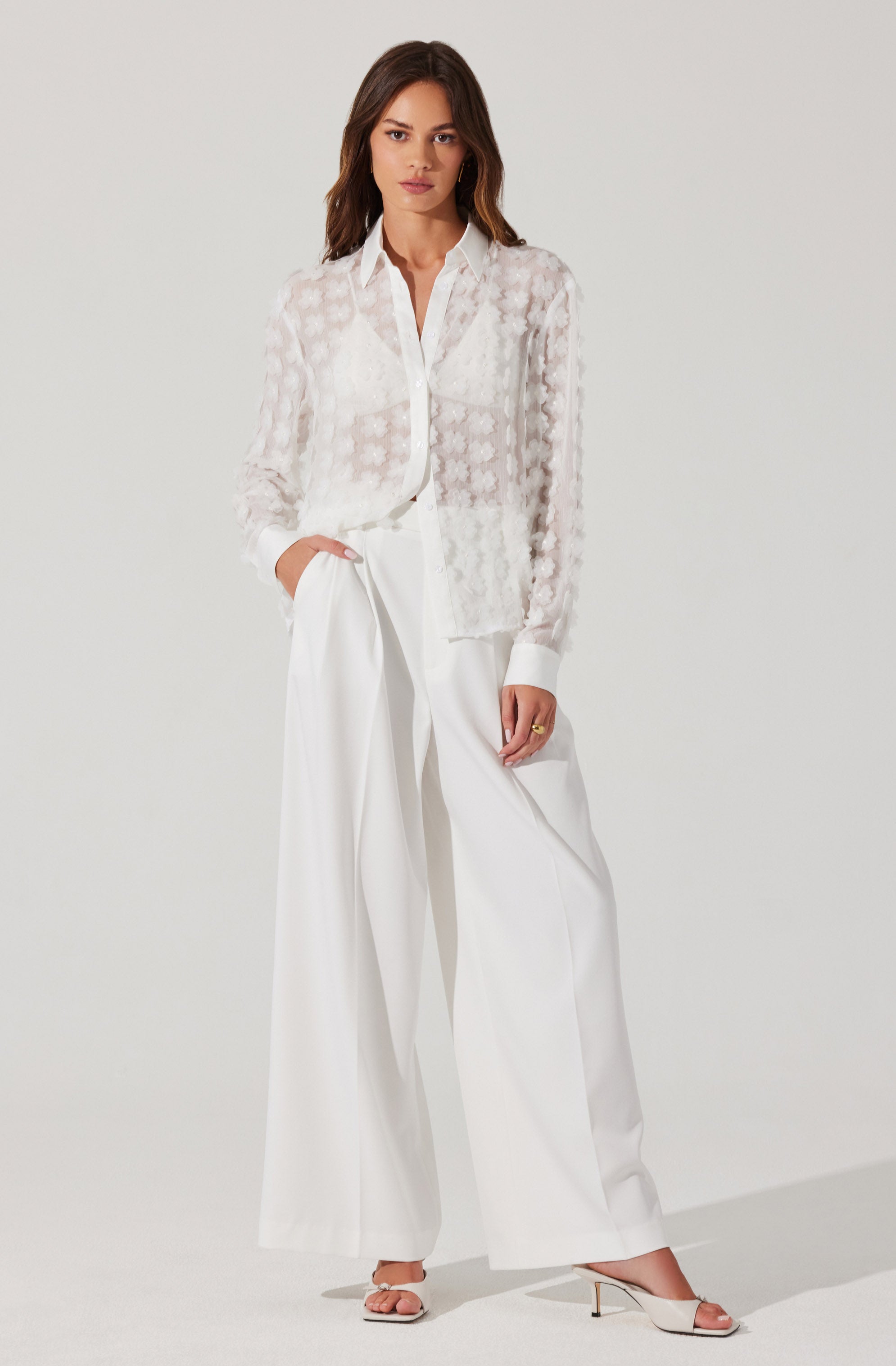A woman is wearing a sheer, long-sleeved white blouse with a subtle floral pattern and a classic shirt collar. The blouse is buttoned down the front, with the top few buttons left open. She pairs the blouse with high-waisted, wide-leg white trousers that have a tailored fit and pleats at the waist. She completes her outfit with white high-heeled sandals featuring a thin strap across the toes and an ankle strap.