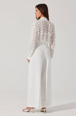 A woman is wearing a sheer, long-sleeved white blouse with textured floral patterns and a collared neckline. She pairs it with high-waisted, wide-leg white trousers and white open-toe high-heeled shoes.