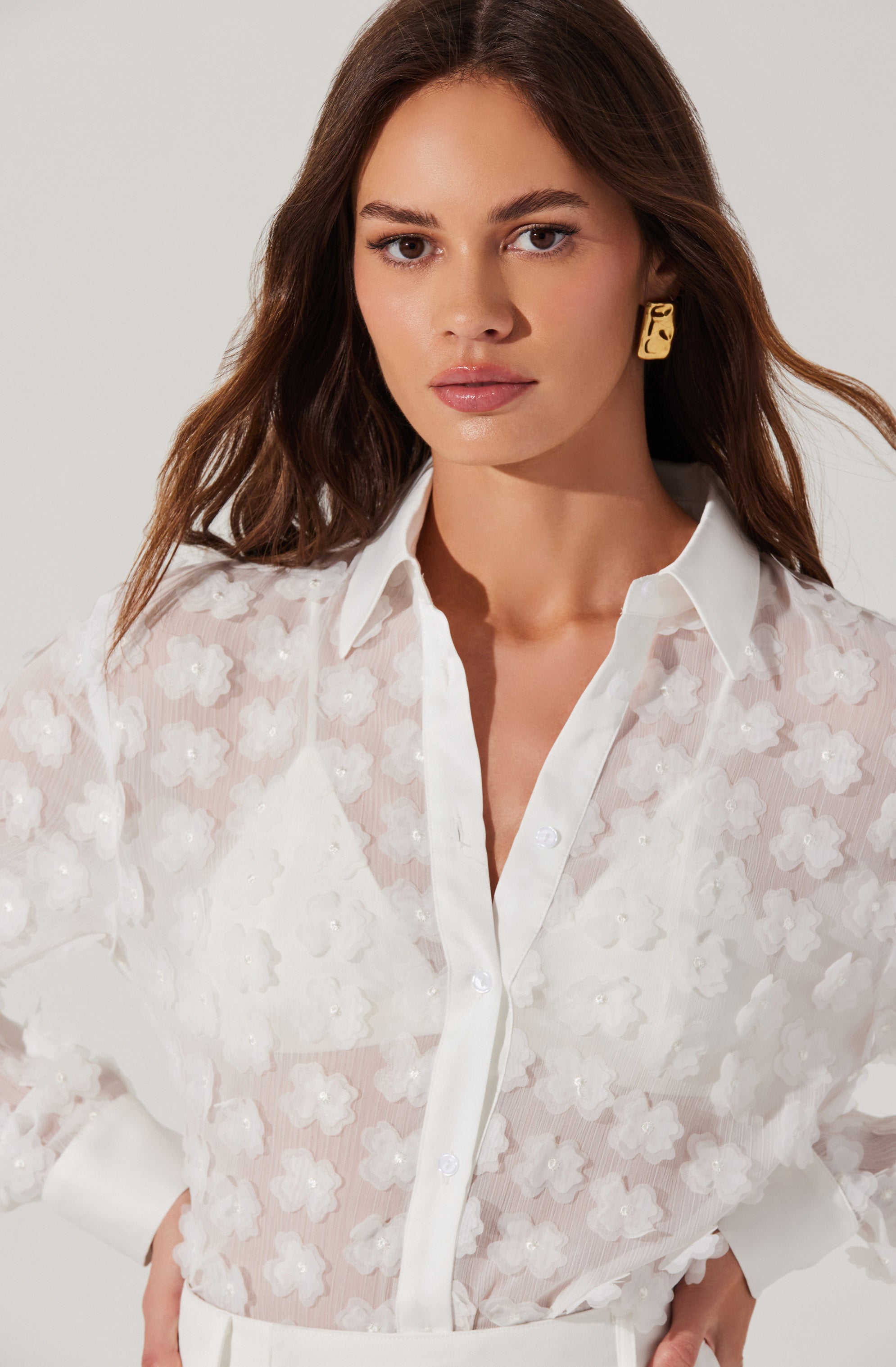 The woman is wearing a sheer white button-up shirt with floral embroidery. The shirt features a classic collar, white buttons down the front, and long sleeves. She has paired it with white bottoms. She is also wearing a large, square-shaped gold earring.
