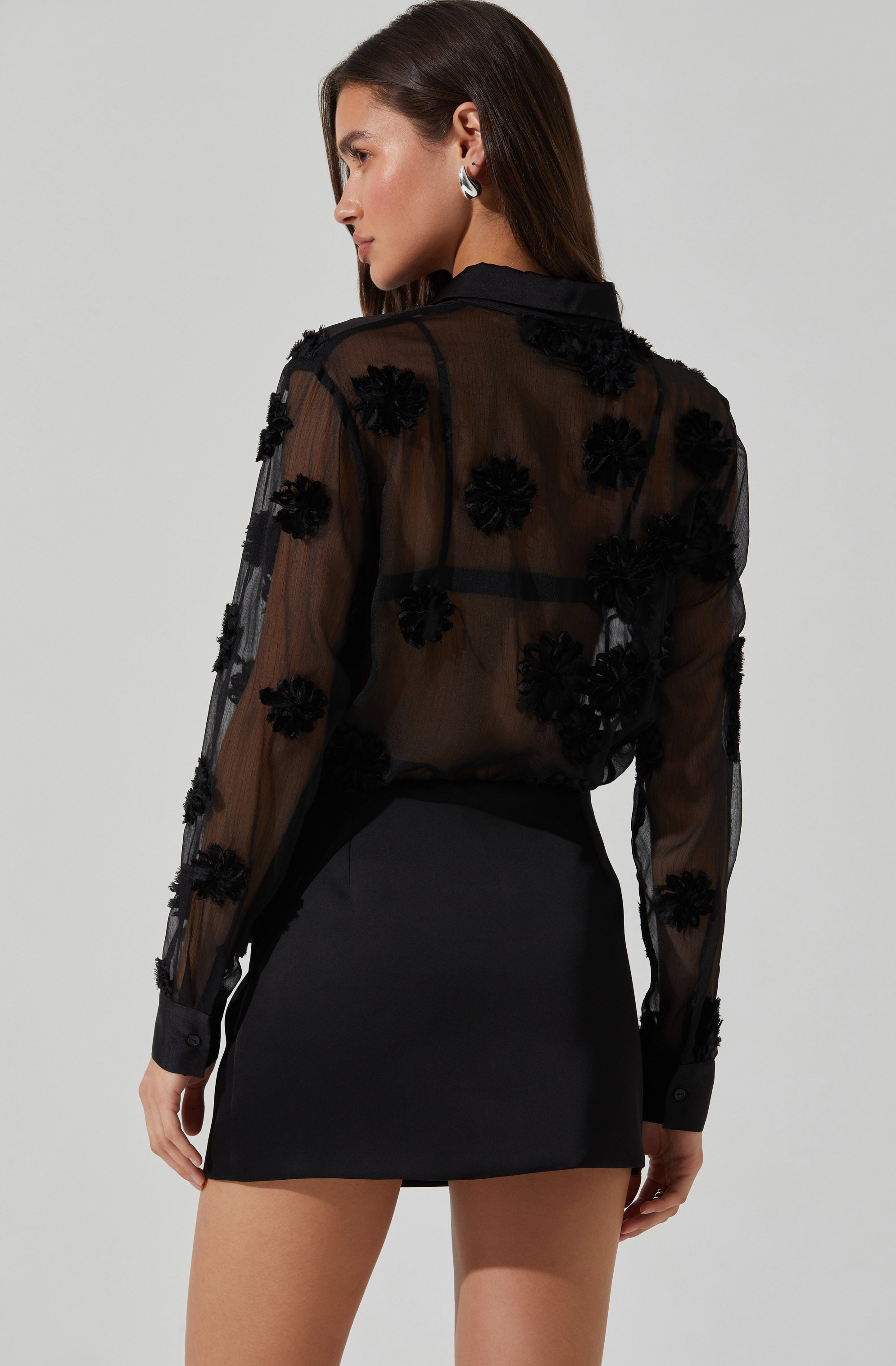 A woman is wearing a sheer black blouse with 3D floral appliqués on the front and sleeves. The blouse has a classic collar and buttoned cuffs. She pairs it with a fitted black high-waisted mini skirt. She has long brown hair and is wearing silver hoop earrings.