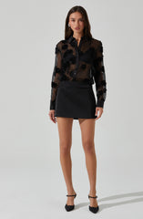 A woman is wearing a black sheer long-sleeve button-up blouse with floral embroidery, paired with a black mini skirt. She is also wearing black high-heeled shoes with ankle straps.