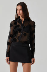 A woman is wearing a sheer black long-sleeve blouse decorated with textured floral appliqués, paired with a high-waisted black mini skirt.