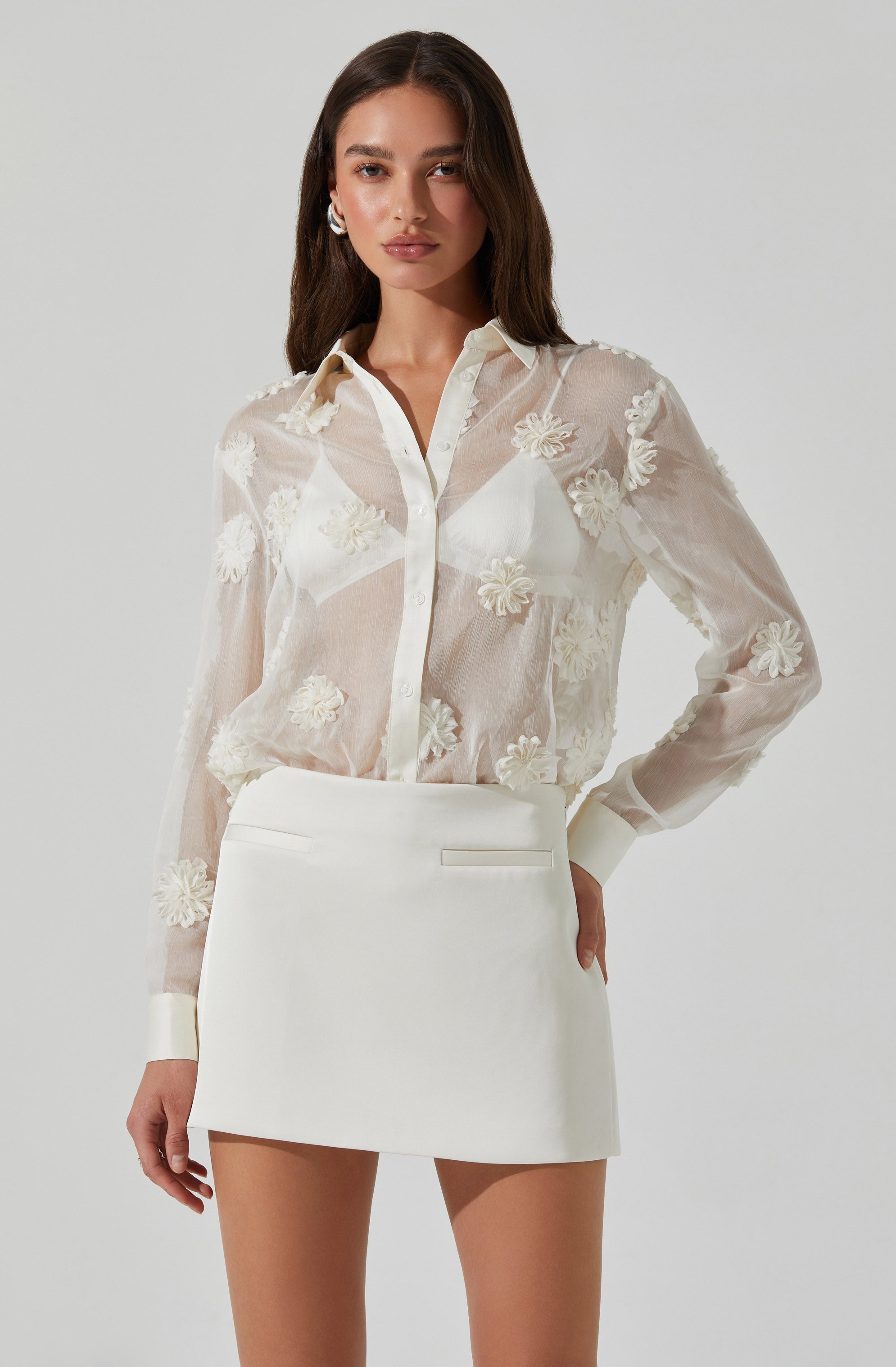 A woman is wearing a sheer, long-sleeved button-up blouse with floral embroidery and a pointed collar, paired with a high-waisted, white mini skirt with a sleek, minimal design.