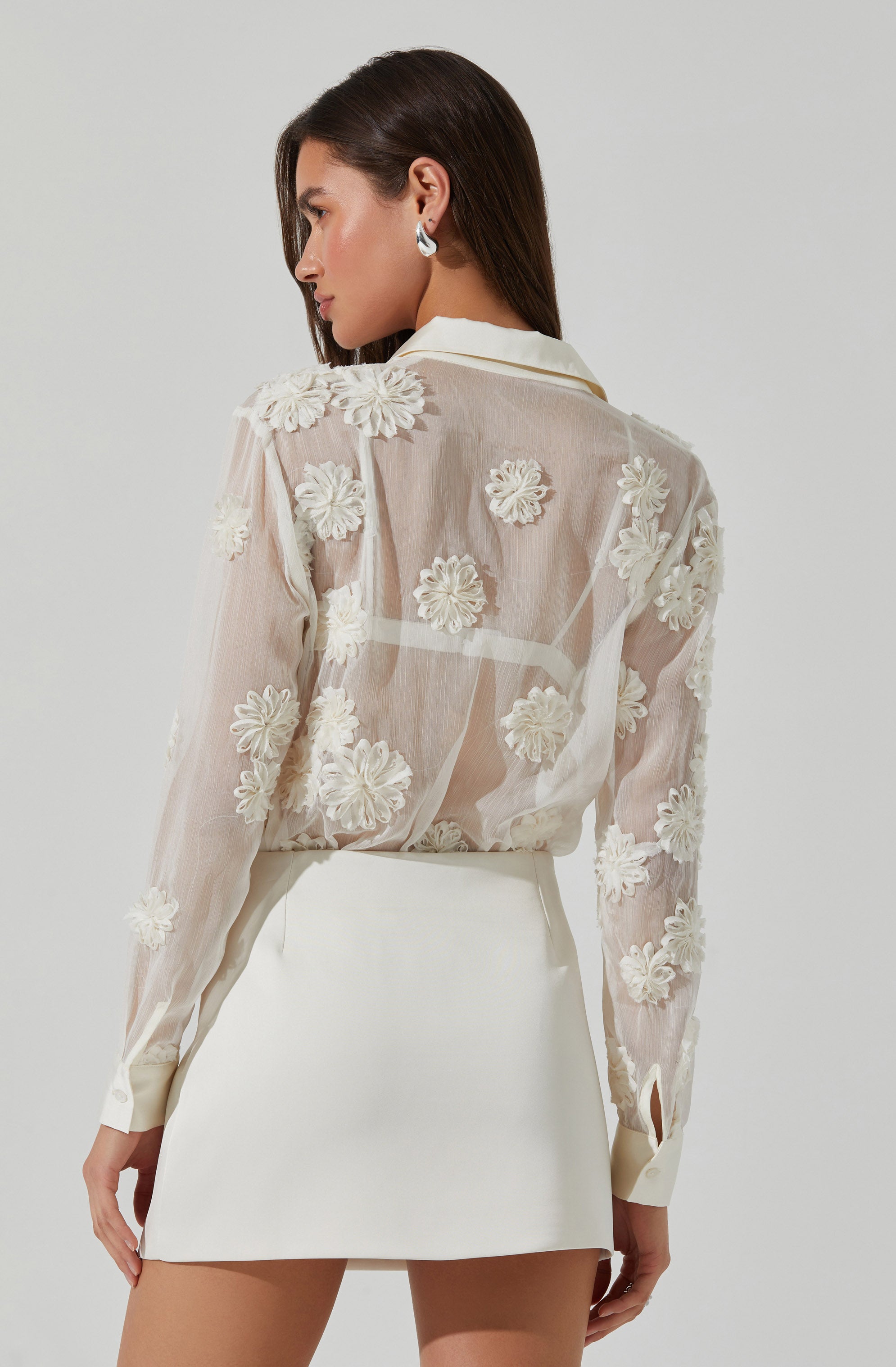 A woman is wearing a sheer, long-sleeved blouse made of lightweight fabric that features 3D floral appliqués throughout. The blouse has a collar and buttoned cuffs at the sleeves. She pairs it with a high-waisted, plain white mini skirt.