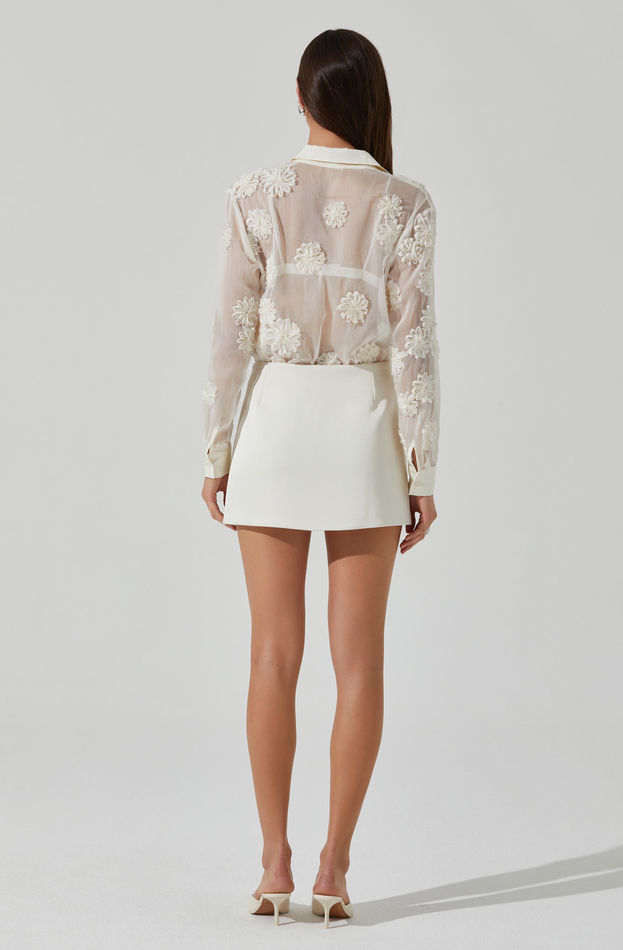 A woman is wearing a sheer, long-sleeved blouse decorated with embroidered floral patterns, paired with a high-waisted, knee-length white skirt. She completes the outfit with light-colored high-heeled shoes.
