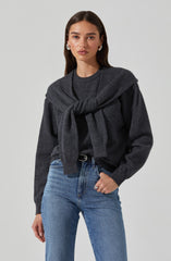 A woman wearing a dark gray sweater with a matching gray sweater tied around her shoulders. She is also wearing light blue high-waisted jeans with a black belt.
