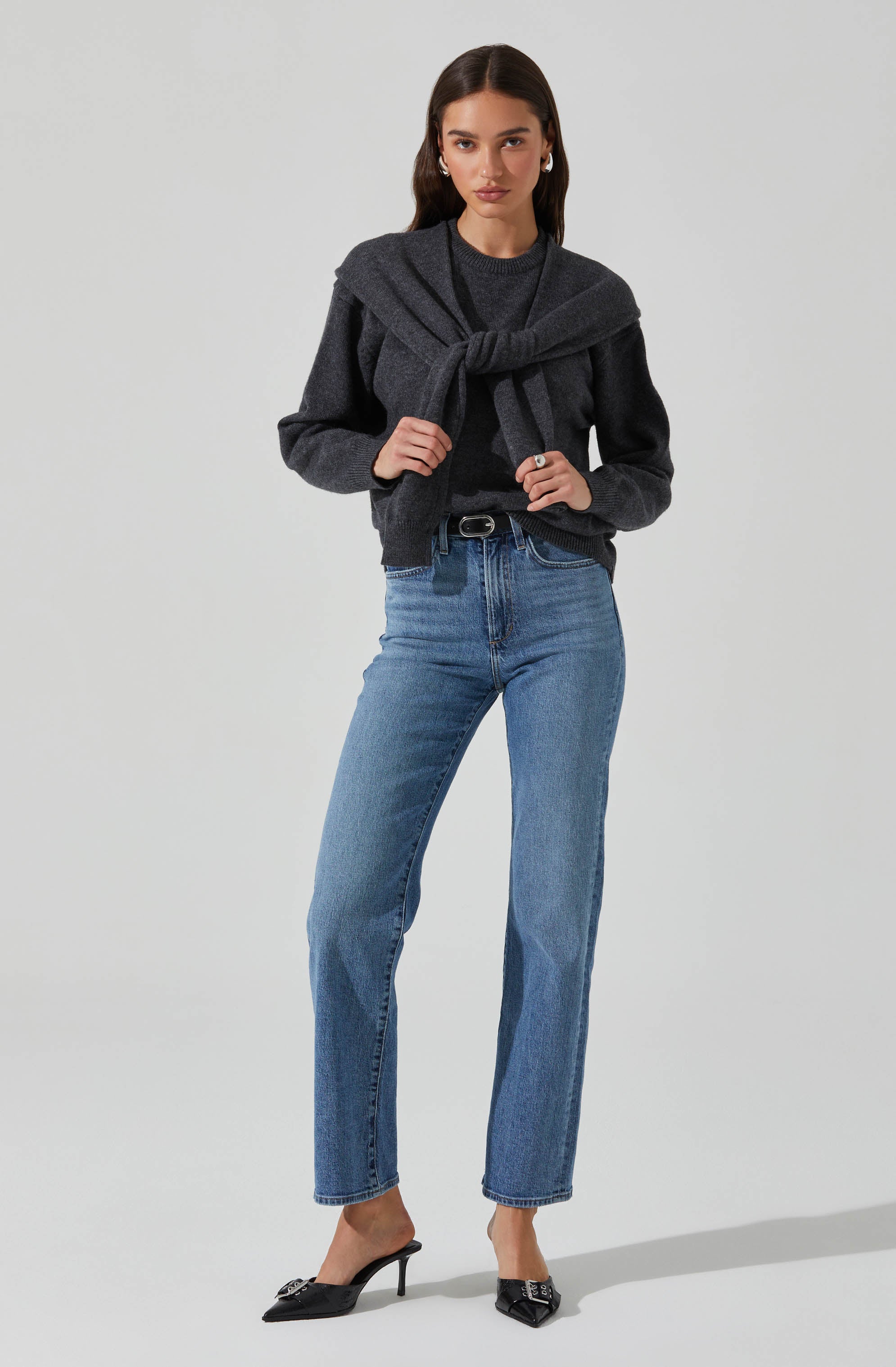 A woman is wearing a dark gray long-sleeved sweater tucked into light blue high-waisted jeans. She has a matching dark gray sweater tied around her shoulders. She is also wearing black pointed-toe high-heeled shoes with decorative embellishments on the front.