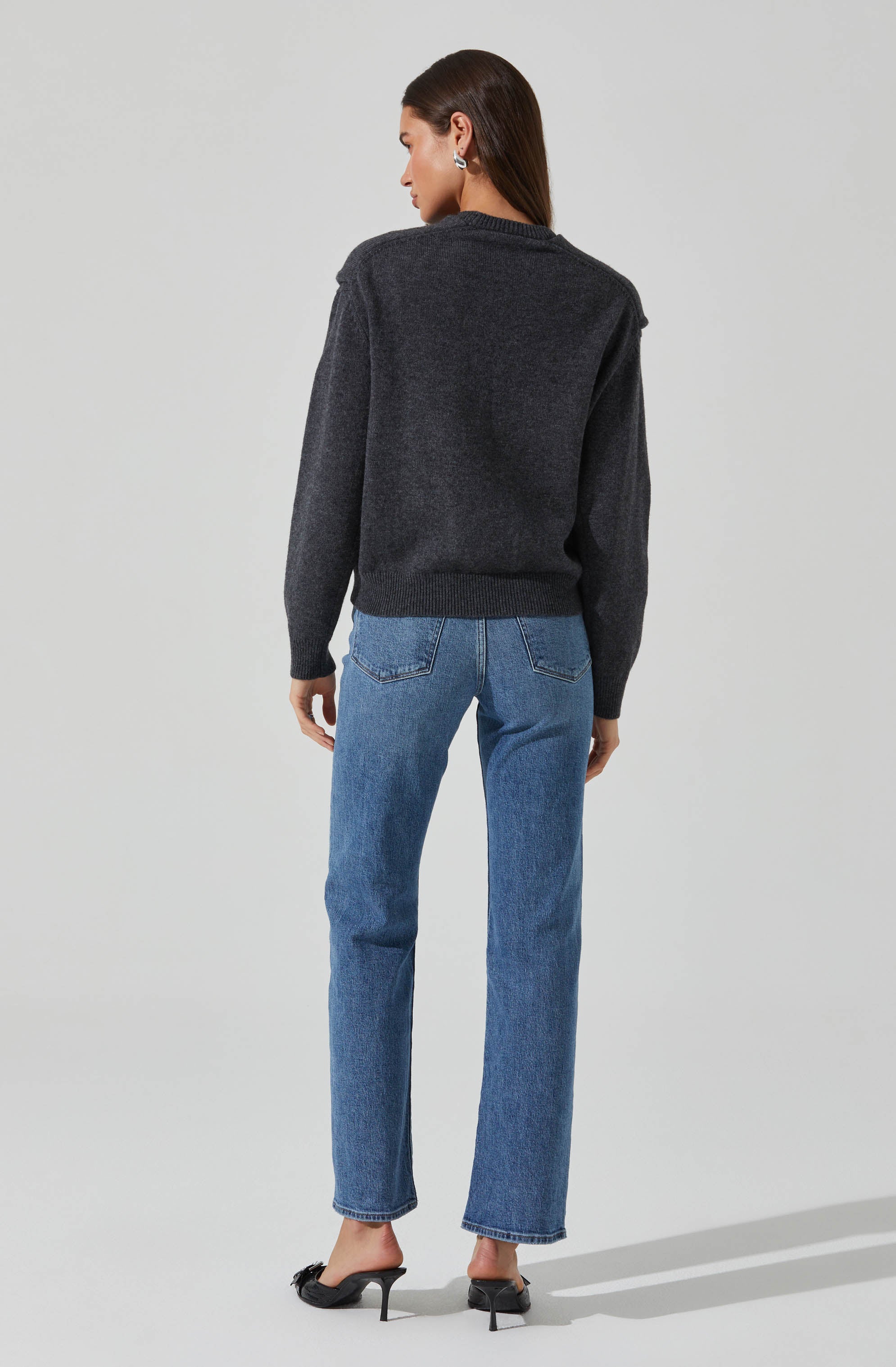 A woman is wearing a dark gray, long-sleeved knit sweater with a relaxed fit and ribbed cuffs and hem. She pairs it with light blue high-waisted jeans that have a slightly relaxed fit and classic back pockets. She completes her outfit with black, pointed-toe high-heeled shoes.