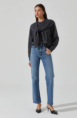 A woman is wearing a dark gray sweater with a matching gray sweater tied around her shoulders. She is also wearing light blue high-waisted jeans and black high-heeled shoes with a pointed toe and bow detail.