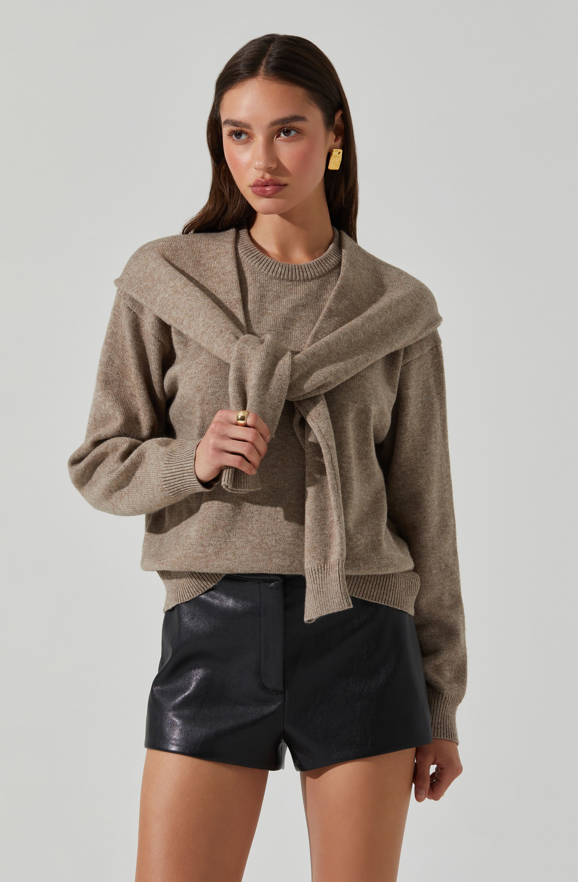 A woman is wearing a beige sweater with a matching scarf draped around her shoulders. She has on gold earrings and a gold ring. She is also wearing black leather shorts.