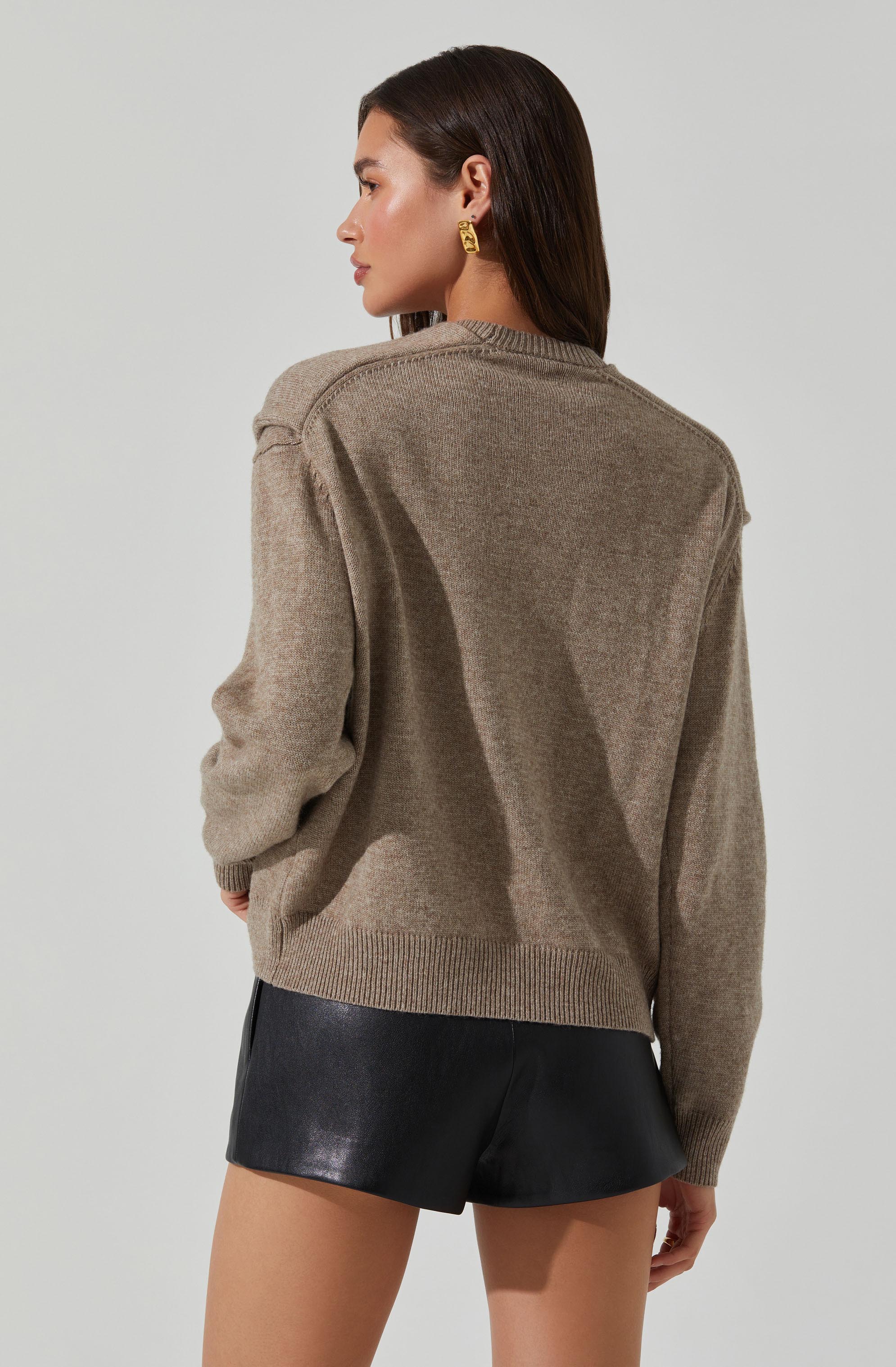 A woman is wearing an oversized, taupe-colored knit sweater with a round neckline and ribbed hem and cuffs. The sweater appears soft and cozy. She pairs it with black leather shorts. She has dark, straight, shoulder-length hair and is accessorizing with gold hoop earrings.