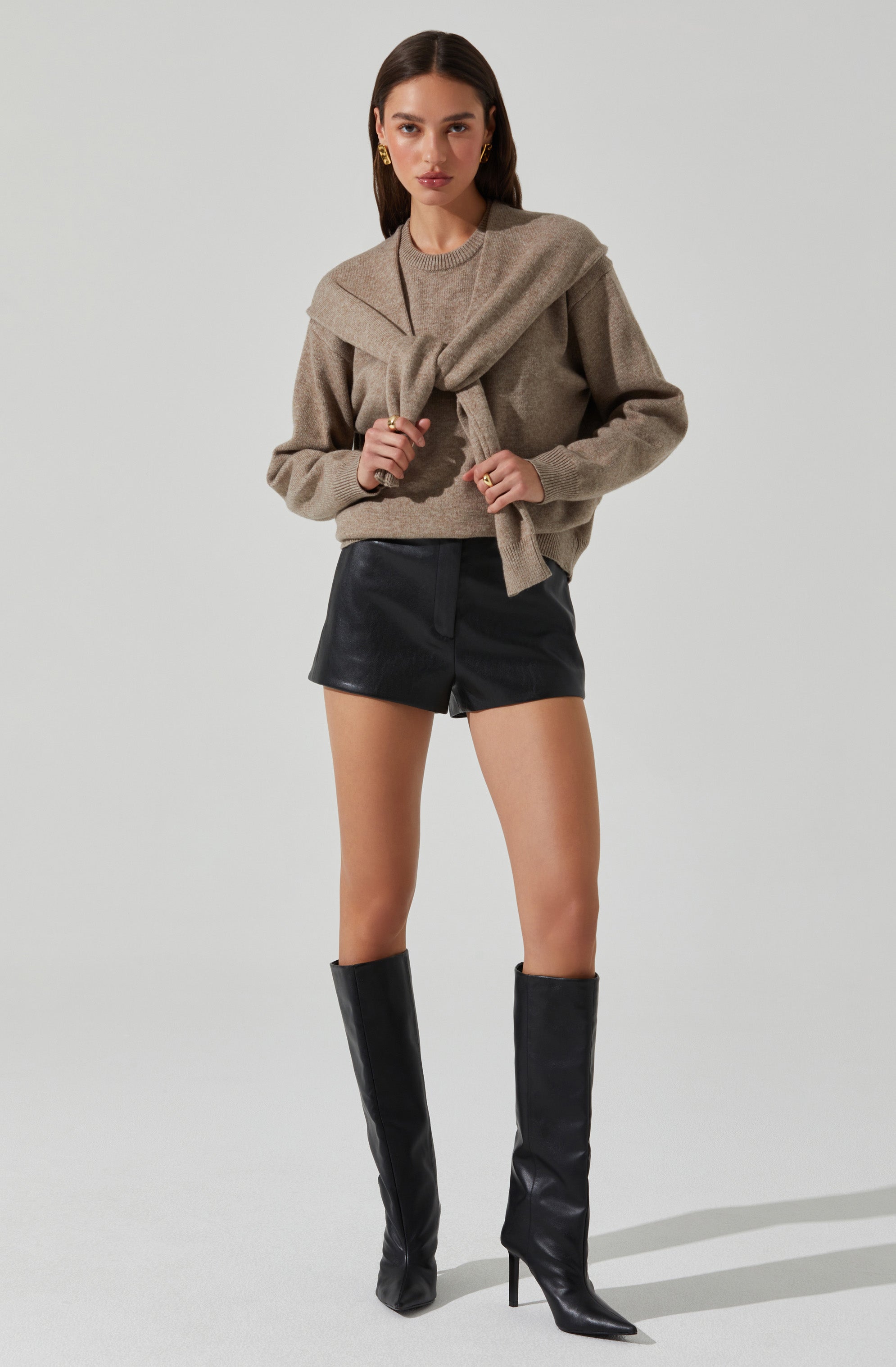 A woman is wearing a taupe-colored oversized sweater with a large collar, layered over a matching sweater tied around her shoulders. She pairs it with high-waisted black leather shorts and tall black leather boots with pointed toes and stiletto heels.