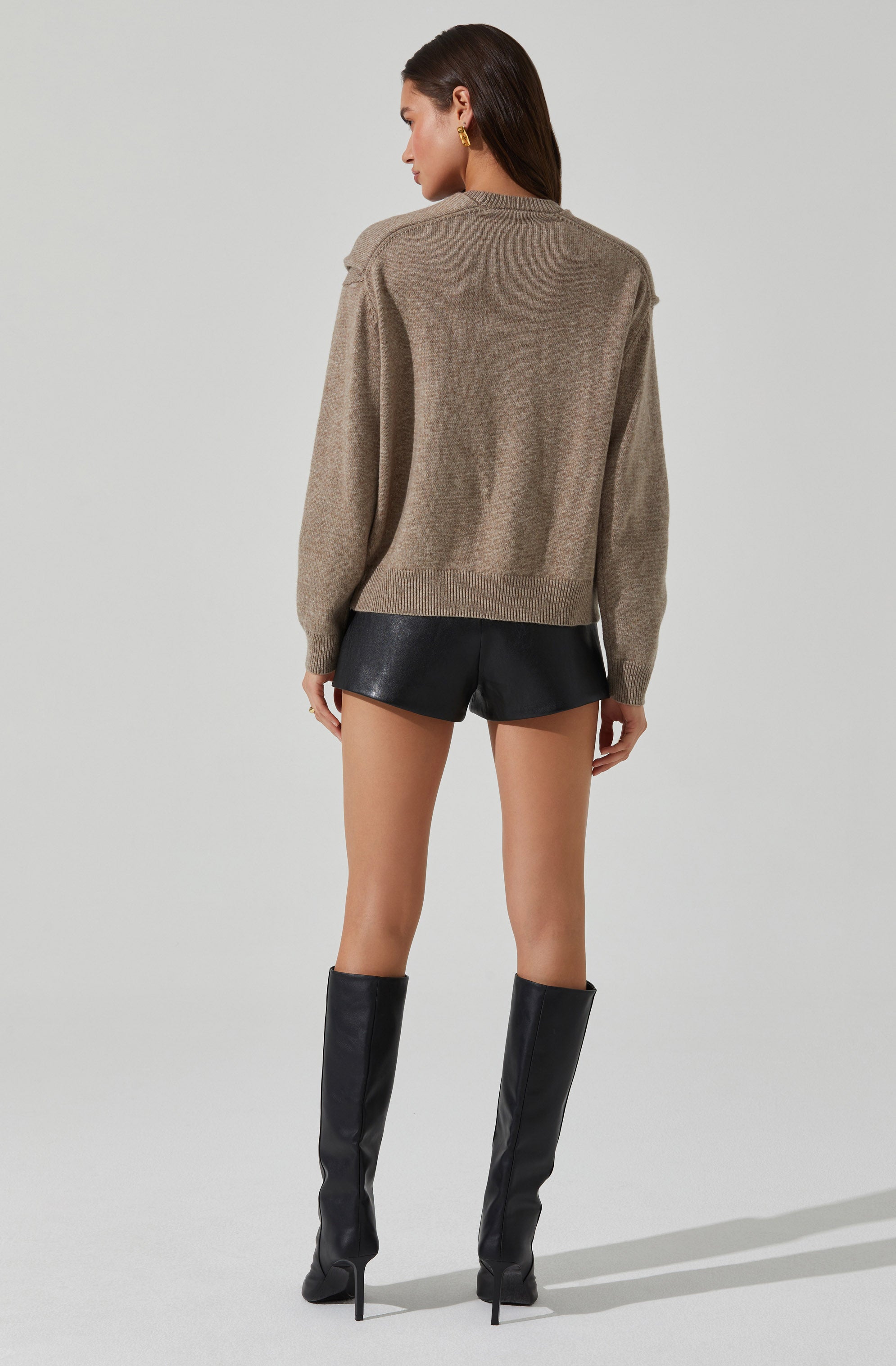 A woman is wearing a loose-fitting, taupe-colored knit sweater with long sleeves and a high neckline. She pairs it with black leather shorts and knee-high black leather boots with high heels.