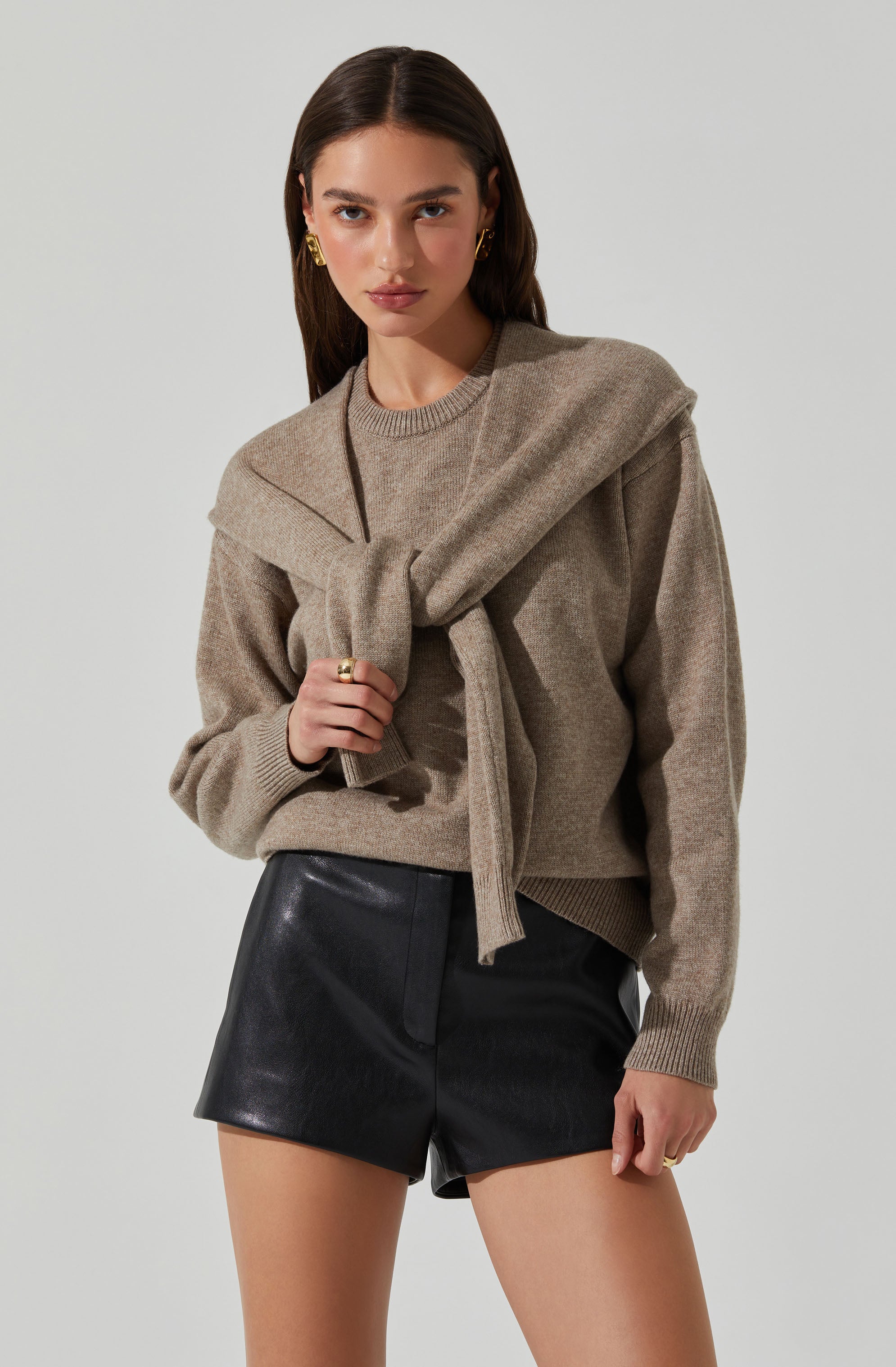 A young woman is wearing a tan knit sweater with a round neckline and long sleeves. She has draped a matching tan sweater around her shoulders, tying the sleeves at the front. She is also wearing high-waisted black leather shorts.