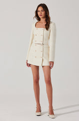 A woman is wearing an elegant, cream-colored two-piece outfit consisting of a textured, long-sleeved blazer with a square neckline and matching high-waisted, double-breasted mini skirt with gold buttons. She is also wearing cream-colored high-heeled shoes.