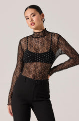 A woman is wearing a long-sleeved, high-neck lace top that is black and sheer, accented with small, white, sparkling dots throughout. She has paired it with a black bra or camisole underneath. She is also wearing high-waisted black pants with a button and zip closure. Her hair is sleek and pulled back, and she has a neutral expression.