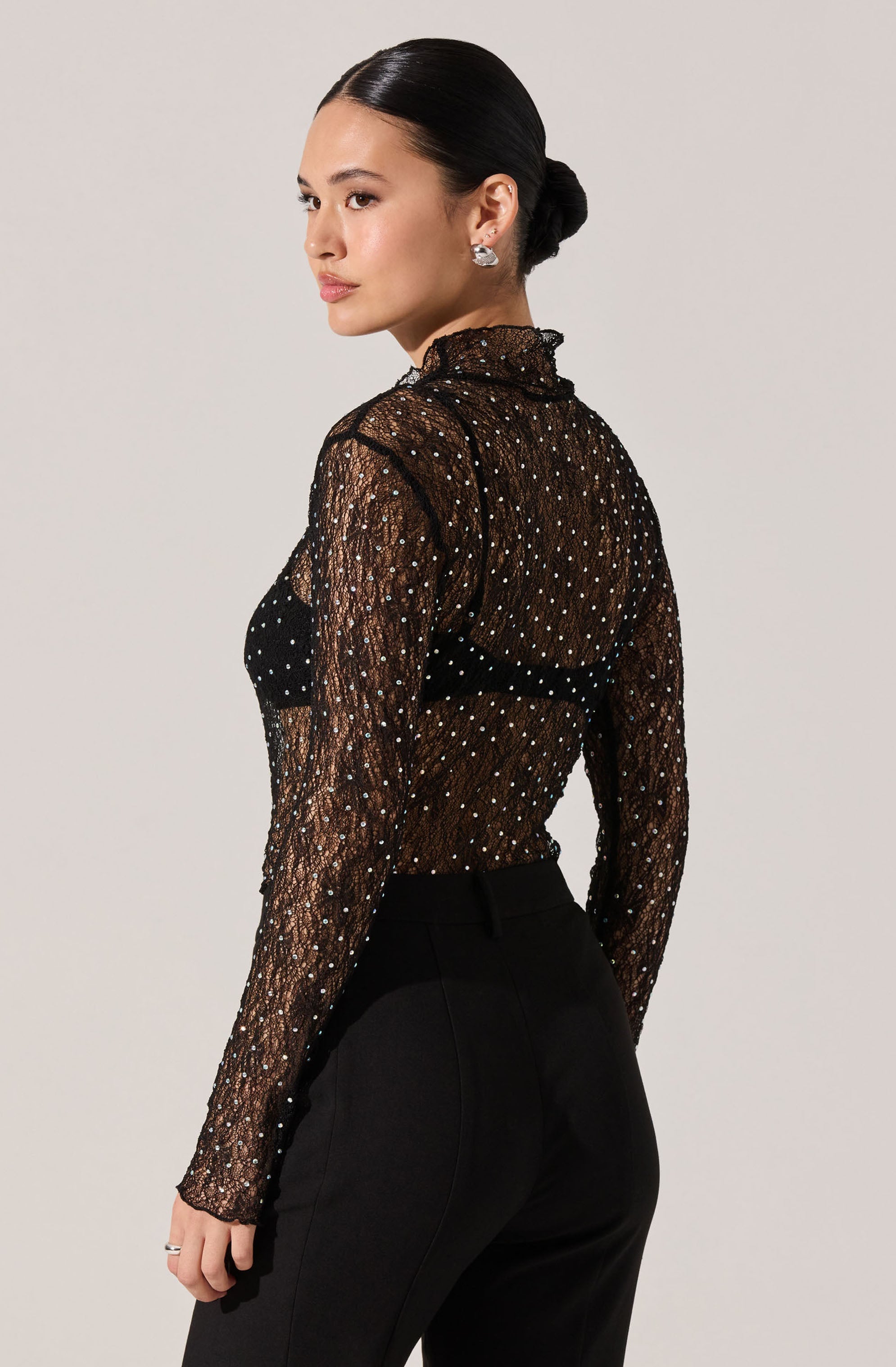 A woman is wearing a sheer black long-sleeve top with a high ruffled collar. The top is decorated with small, shiny, white polka dots. She pairs it with high-waisted black pants.