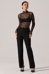 A woman is wearing a sheer black long-sleeve top with a high neckline, decorated with small, shiny embellishments. Underneath, she has a black bralette that is visible through the sheer fabric. She is also wearing high-waisted black trousers with a straight-leg fit, featuring a zipper and button closure, and small zippers at the hem. She completes the look with black heels that have a decorative buckle on the front.